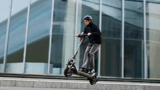 Helmeted rider jumping an electric scooter in front of a modern glass building.