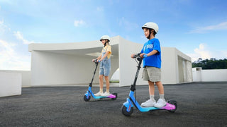 Two children wearing helmets riding NAVEE K100 Kids Electric Scooters on a paved area.